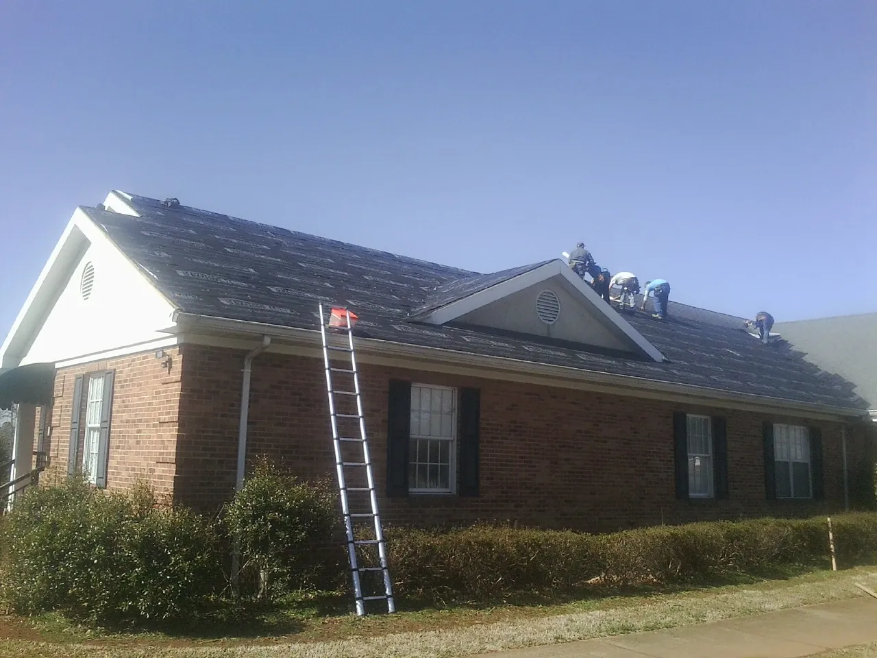 Crew tearing shingles off a brick ranch roof with a ladder set — Emergency Roof Repair work by our crew in Linkwood